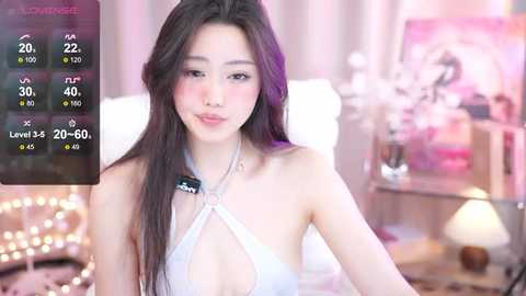 Media: Video of a young Asian woman with long dark hair, wearing a revealing white halter top, in a softly lit room with pastel decor.