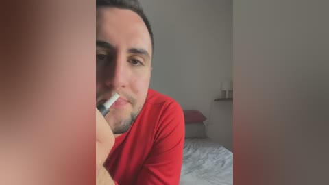 Media: Video of a man with short dark hair, wearing a red shirt, lying on a bed, holding a white toothbrush in his mouth, with blurred background.