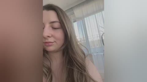 Media: Video of a young Caucasian woman with long, straight, light brown hair, light skin, and closed eyes, standing in a dimly lit room with white curtains.