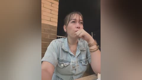 Media: Video of a young woman with dark hair in a brick building, wearing a denim shirt, with a contemplative expression, looking downward.