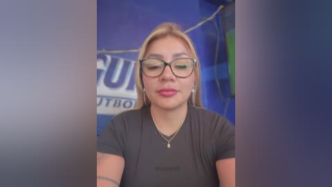 Media: Video of a young woman with fair skin, light blonde hair, and black-rimmed glasses, wearing a gray t-shirt, standing in front of a blue wall with blurred text.