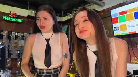 Media: Video of two young women in a bar, dressed in school uniforms with black ties, smiling, in a dimly lit room with neon signs and a TV screen.