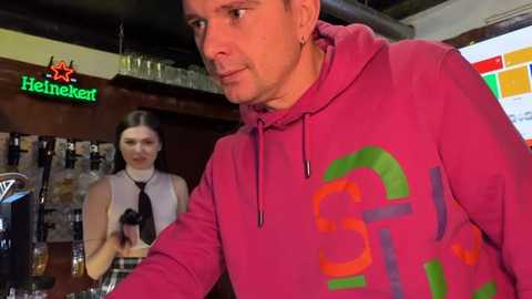 Media: A video of a bar scene featuring a man in a red hoodie with a graphic design, pouring a beer, and a bartender in a white sleeveless top, both looking toward the camera.