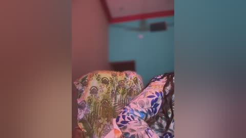 Media: A video of a bed with a person partially visible beneath colorful, patterned bed covers. The background features a teal wall with red trim, and a ceiling fan.