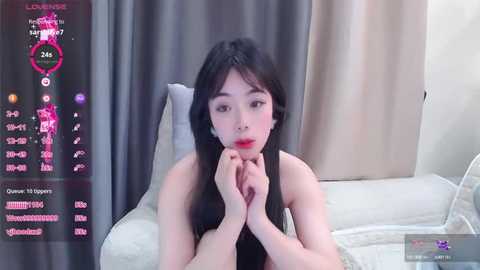 Media: A video of an East Asian woman with long black hair, fair skin, and red lipstick, wearing a black top, sitting on a beige couch in a modern room.