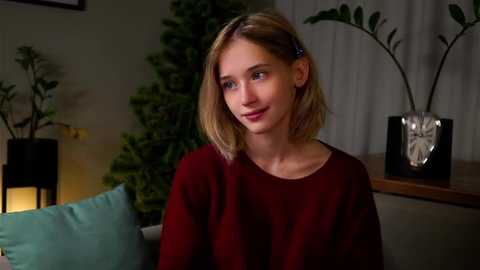 Media: Video of a young Caucasian woman with light blonde hair, wearing a red sweater, sitting on a couch, surrounded by indoor plants, dimly lit room.