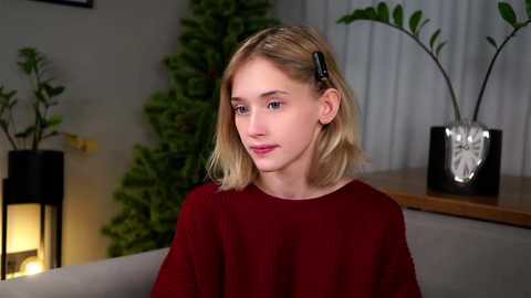 Media: Video of a young blonde girl with shoulder-length hair, wearing a red shirt, sitting indoors with potted plants and a metallic vase on a wooden shelf in the background.