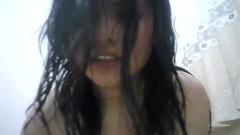 Media: Video of a woman with long, wet, dark hair covering her face, partially visible mouth, and a white lace curtain in the background.