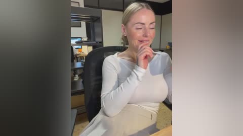 Media: Video of a blonde Caucasian woman in a light grey sweater, sitting at a desk with a black chair, smiling, fingers touching her chin. Background shows a modern office with dark cabinets and a kitchen area.