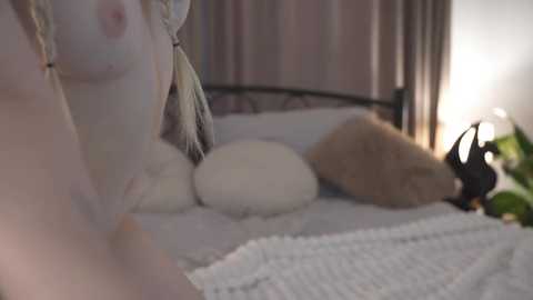 Media: Video of a partially nude woman with pale skin, medium-sized breasts, and braided hair, lying on a bed with white and beige bedding. The background features curtains and a lamp.