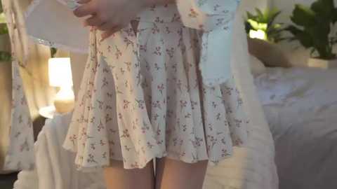 Media: Video of a woman in a white, floral-patterned dress, adjusting the hem. Background features a bed with white sheets, a potted plant, and a lamp. The setting appears to be a cozy, well-lit bedroom.