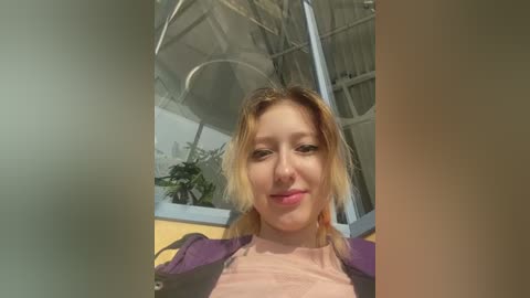 Media: Video of a young Caucasian woman with light blonde hair, fair skin, and light makeup, wearing a purple jacket over a light pink shirt, smiling inside a greenhouse with potted plants and a metal frame ceiling.