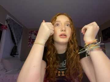 Media: Video of a young, fair-skinned, curly-haired woman with glasses, wearing a black shirt, bracelets, and a necklace, striking a boxing pose in a dimly lit bedroom.