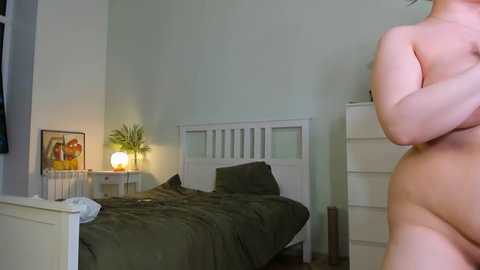 Media: Video of a modestly furnished bedroom with a white bed, olive-green bedding, and a white nightstand with a framed picture and lamp. A nude person stands in the right foreground, partially visible.