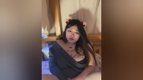 Media: Video of an Asian woman with long black hair, wearing a black off-shoulder top and a red bow, sitting on a bed with floral-patterned sheets, in a dimly lit room.