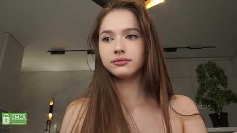 Media: Video of a young woman with fair skin and long brown hair, wearing a beige top, indoors with a modern, minimalist background featuring a green plant and white walls.