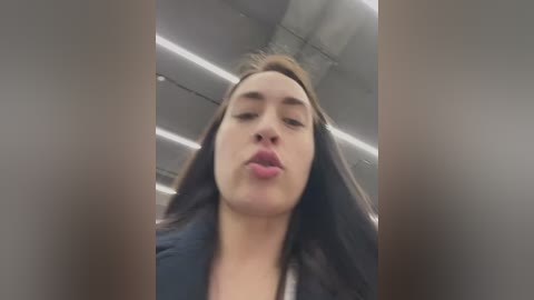 Media: Video of a woman with light skin, long dark hair, and full lips, blowing a kiss. She wears a dark top. Background features a modern, industrial ceiling with visible ductwork.