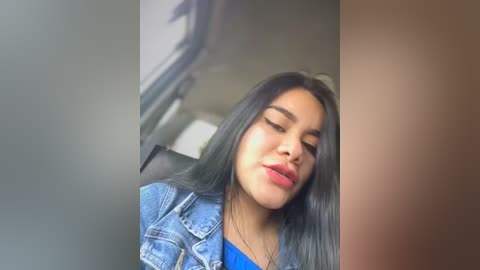 Media: Video of a young Latina woman with long black hair, wearing a blue denim jacket, red lipstick, and a closed-mouth smile. Background shows a blurred car interior.