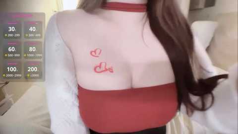 Media: Video of a woman with fair skin and long dark hair, wearing a red choker and a sheer white top revealing large breasts with red heart tattoos, in a bedroom with a bed and a digital health tracker screen.