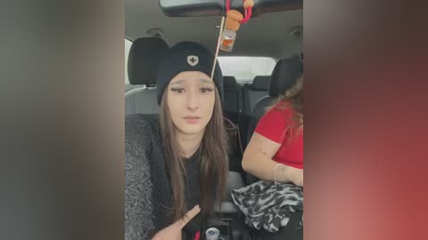 Media: Video of a young woman with long brown hair, wearing a black beanie and a grey sweater, seated in a car, looking downcast.