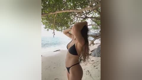 Media: Video of a slender, light-skinned woman with long, dark hair in a black bikini, standing under a tree on a tropical beach with turquoise waters and rocky outcrops in the background.