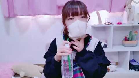 Media: Video of an Asian woman with brown hair wearing a white surgical mask, black jacket, and green plaid shirt, spraying disinfectant in a pastel-colored room with white shelves, plush toys, and a pink curtain.