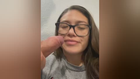 Media: A video showing a young woman with glasses, light skin, and long brown hair, smiling as she holds a man's erect penis near her lips.