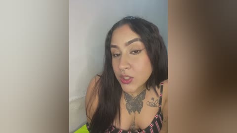 Media: A video of a Latina woman with long black hair, wearing a black and pink striped top, showcasing a detailed butterfly tattoo on her chest and an additional tattoo on her neck. She has a medium skin tone and is smiling slightly.