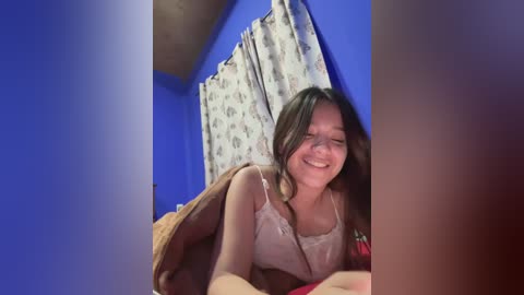 Media: A video of a young woman with long brown hair, wearing a white lace top, smiling while lying on a bed with a red blanket. The background features blue walls and patterned curtains.