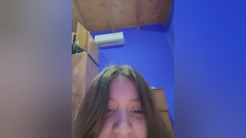 Media: A video of a young woman with long, straight brown hair, captured from a low angle, looking down. The room features a wooden ceiling, blue walls, and a white air conditioner.