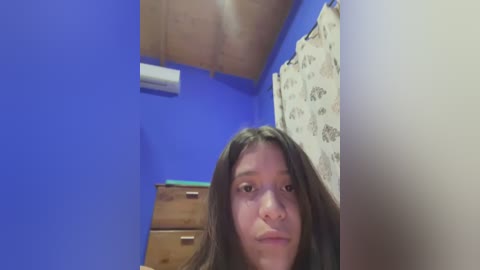 Media: Video of a young Latina woman with long dark hair, standing in a small, brightly lit room with blue walls. She has a neutral expression. The room features a wooden dresser, an air conditioner, and a patterned shower curtain.