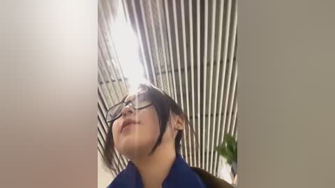 Media: A video of a young Asian woman with glasses, fair skin, and short dark hair, smiling upward. She stands in a modern, brightly lit office with vertical metal blinds and a potted plant in the background.