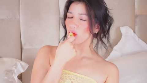 Media: Video of an Asian woman with fair skin and black hair, wearing a yellow lace dress, biting her finger with closed eyes, seated on a bed with white pillows and a beige headboard in a softly lit room.