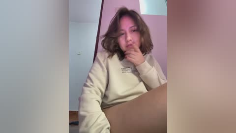 Media: Video of a young woman with shoulder-length, wavy brown hair, wearing a beige hoodie, sitting on the floor, with a reflective mirror partially obscuring her face. Background shows a minimalist room with white walls and a wooden floor.