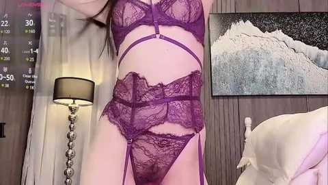 Media: Video of a woman in purple lace lingerie standing in a bedroom with a bed, lamp, and mountain painting on the wall.