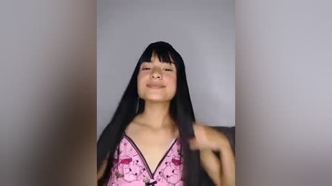 Media: Video of a smiling East Asian woman with long black hair and bangs, wearing a pink bra with a floral pattern, standing in a plain, white-walled room.