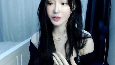 Media: Video of an East Asian woman with long, dark hair, wearing a black off-shoulder top, standing in a bathroom. She has a pale complexion and a subtle, serene expression.