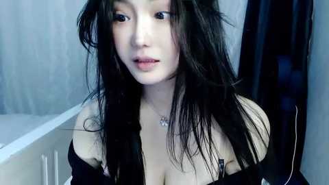 Media: A video of an East Asian woman with long, wet, black hair, fair skin, and full lips, wearing a black off-shoulder top, standing indoors with blurred background.