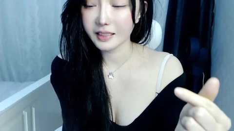 Media: Video of a young Asian woman with long black hair, fair skin, and a slender physique. She wears a black top with a white strap, and a silver necklace. Background shows a modern bathroom with a white bathtub and a gray tiled wall.