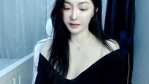 Media: Video of a young Asian woman with long black hair, wearing a black off-shoulder top, seated indoors. She has fair skin, a slender build, and a small, delicate necklace. The background features a white bathtub and a shower curtain.