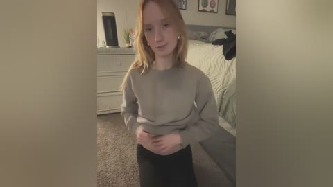 Media: Video of a young woman with fair skin and long blonde hair, wearing a grey sweater and black pants, standing in a cozy bedroom with beige carpet, a white dresser, and a neatly made bed in the background.