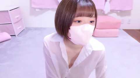 Media: Video of a young Asian woman with short brown hair and a white surgical mask, wearing a white shirt, standing in a pastel-colored, minimalist room with pink furniture and drawers.