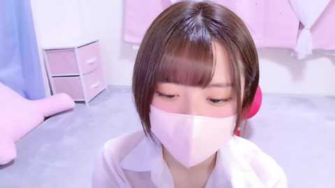 Media: Video of a young Asian woman with short brown hair, wearing a white shirt and pink face mask, sitting on a bed in a pastel-pink room.