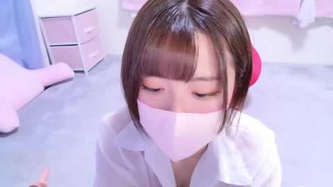 Media: Video of an Asian woman with straight brown hair, wearing a white surgical mask and white shirt, kneeling in a pastel-colored room with a pink bed and white furniture.