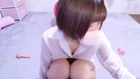 Media: Video of a young Asian woman with straight, shoulder-length brown hair, wearing a white blouse and black panties, kneeling on a light gray carpet, with a pink vibrator nearby.