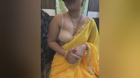 Media: Video of a South Asian woman in a yellow sari, revealing a beige lace bra. She has medium skin tone and wears a gold necklace. Background includes a dark patterned wall and a green cloth.