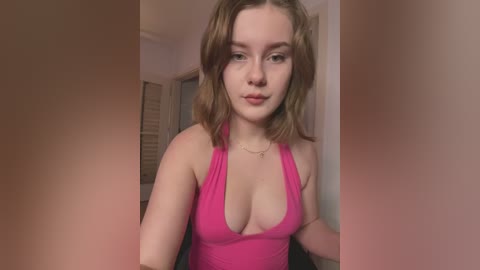 Media: Video of a young Caucasian woman with light skin, wavy brown hair, and pink tank top, showing cleavage, standing indoors with beige walls and closed shutters in the background.