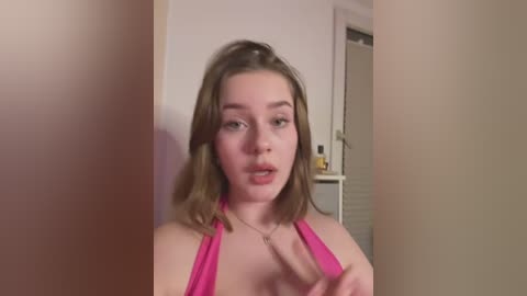 Media: A video of a young Caucasian woman with light skin and shoulder-length brown hair, wearing a pink halter top, standing in a dimly lit room with beige walls. She has a neutral expression.