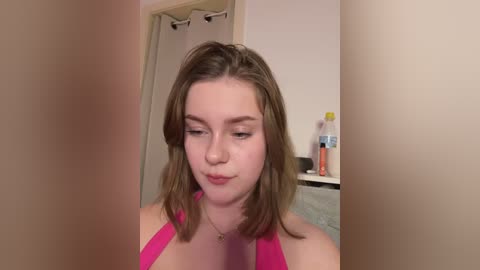 Media: Video of a young Caucasian woman with shoulder-length light brown hair, wearing a pink halter top, standing in a bathroom with beige walls, a shower curtain, and toiletries on a shelf.