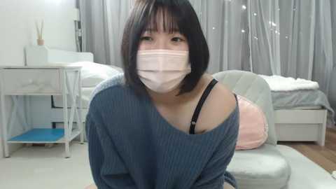 Media: Video of an Asian woman with short black hair, wearing a blue sweater, face mask, and black bra straps, sitting on a bed in a softly lit, modern bedroom with light gray curtains and white furniture.